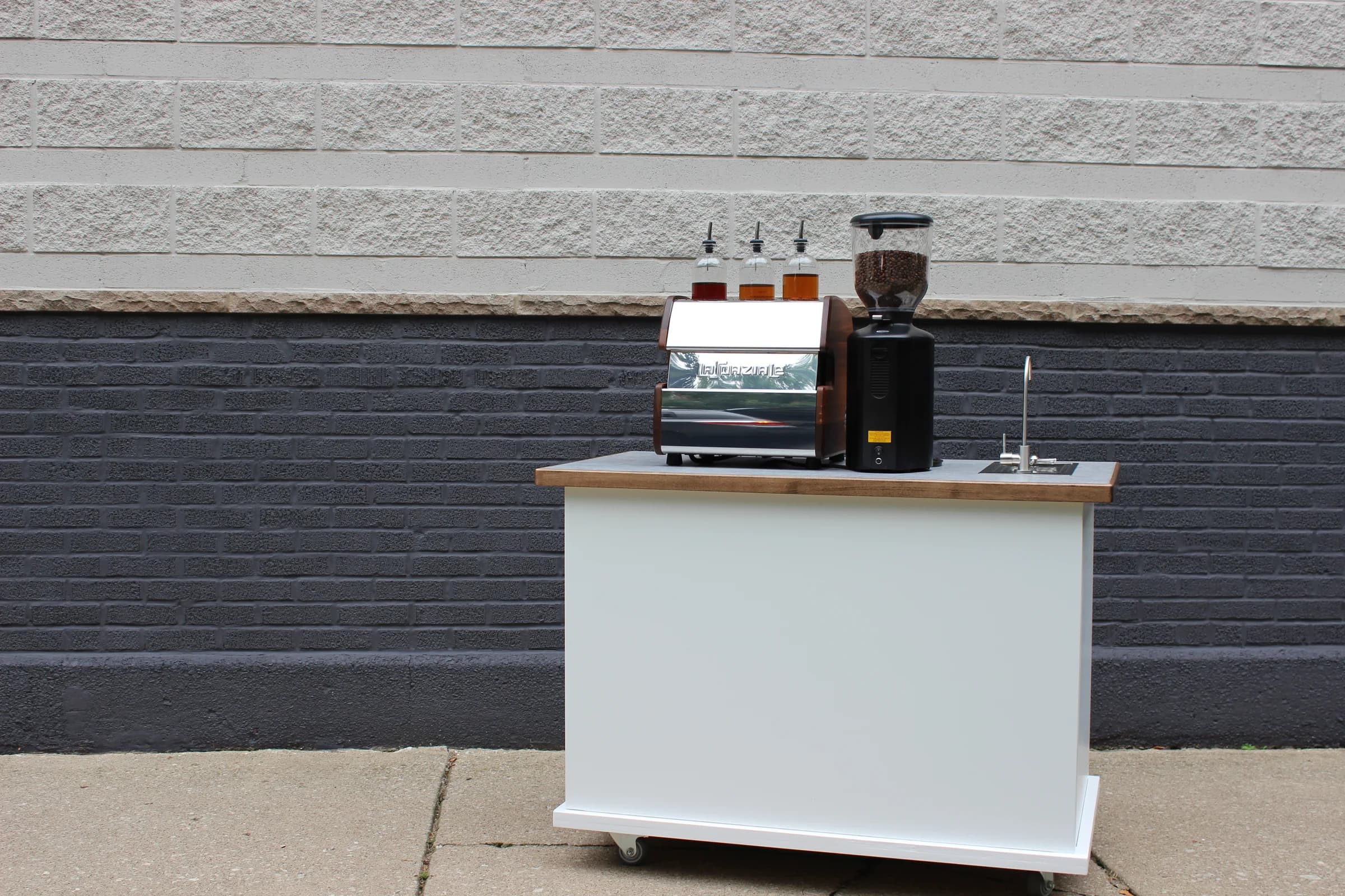 The smaller cart with syrup bottles, espresso machine, and grinder against a cream and slate-blue brick wall