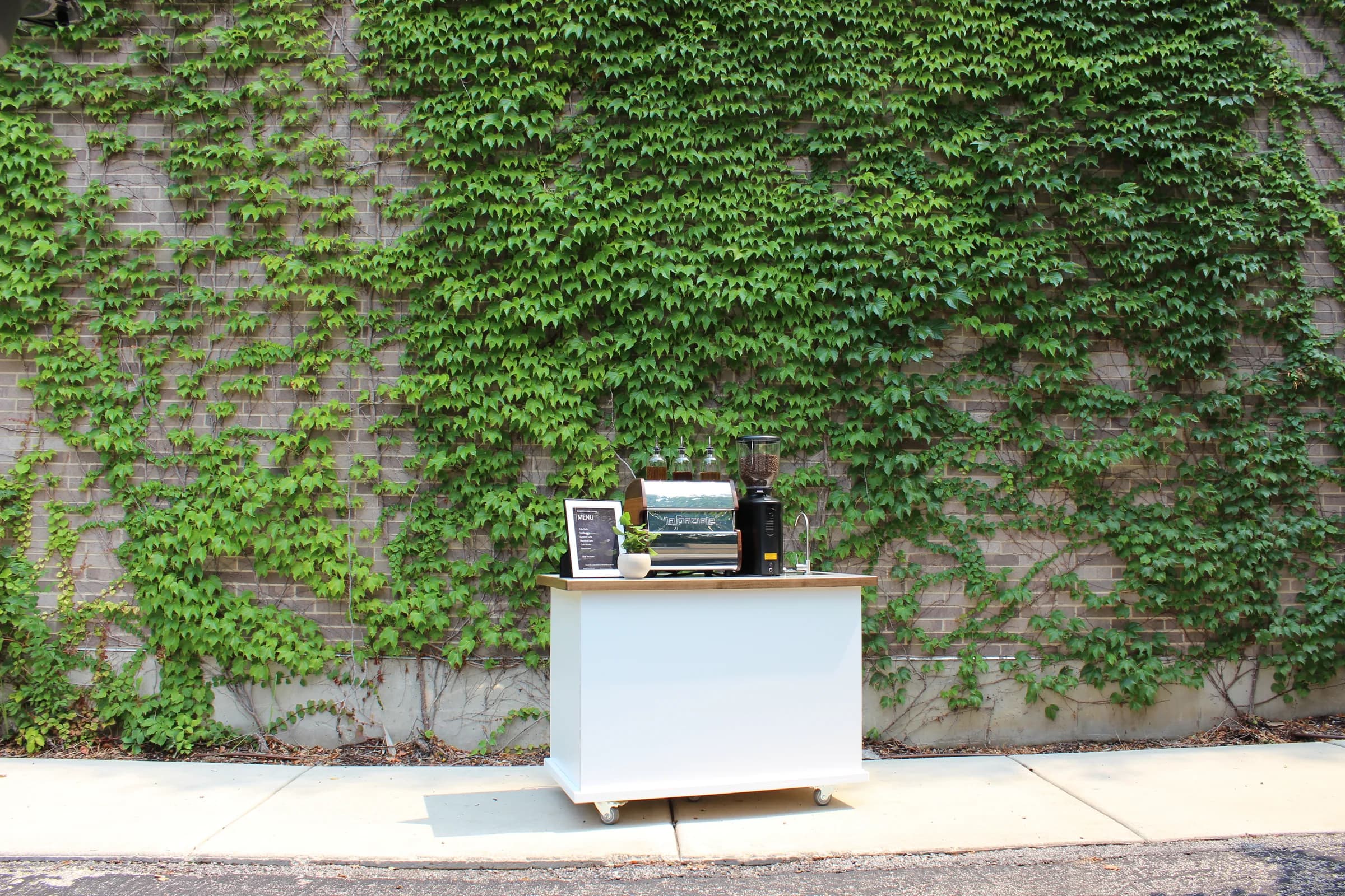 The smaller Wooden Cow Coffee espresso cart, white with a walnut counter, centered against a tall ivy-covered brick wall